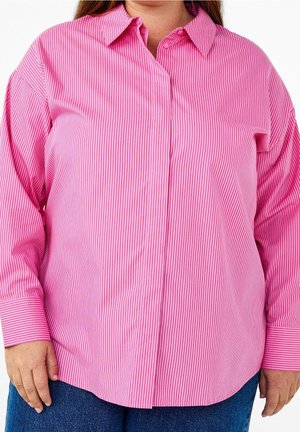 Osoba ubrana w długim rękawem różową koszulę z cienkimi pionowymi paskami i niebieskie dżinsy, ręce swobodnie opuszczone wzdłuż ciała, na białym tle.