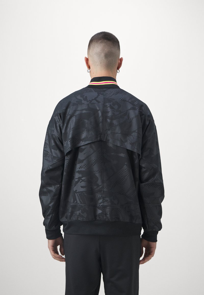 Persona con capelli corti e orecchini a cerchio, che indossa una giacca nera a motivi con un collo a strisce neon, vista da dietro su uno sfondo semplice.