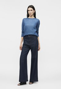 Ženska stoji oblečena v modro majico z dolgimi rokavi, temne široke kavbojke, črne odprte salonarje in uhane v obliki obročev na enobarvnem ozadju.