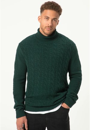 Suéter de cuello alto de punto en verde oscuro con patrón de trenzas, puños y dobladillo acanalados, llevado sobre una camisa blanca, contra un fondo claro.