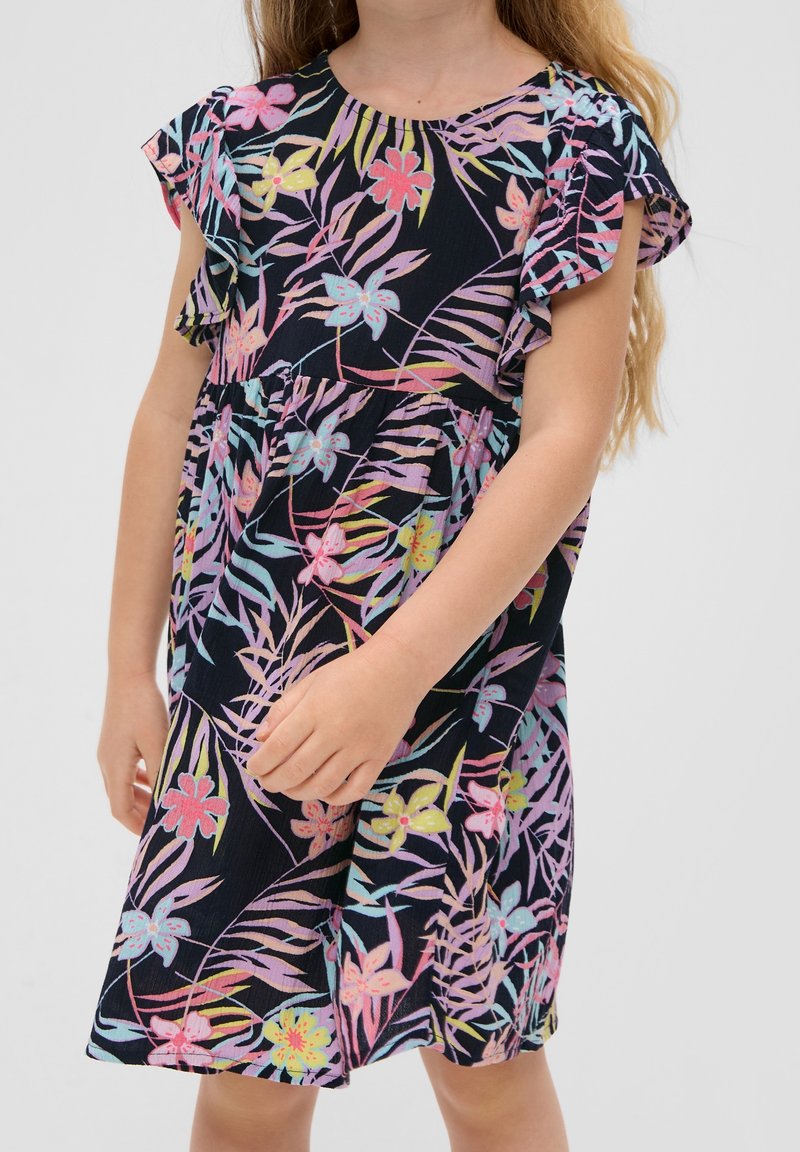 Fille portant une robe sans manches bleu marine avec des imprimés floraux et de feuilles colorés, debout devant un fond clair uni.