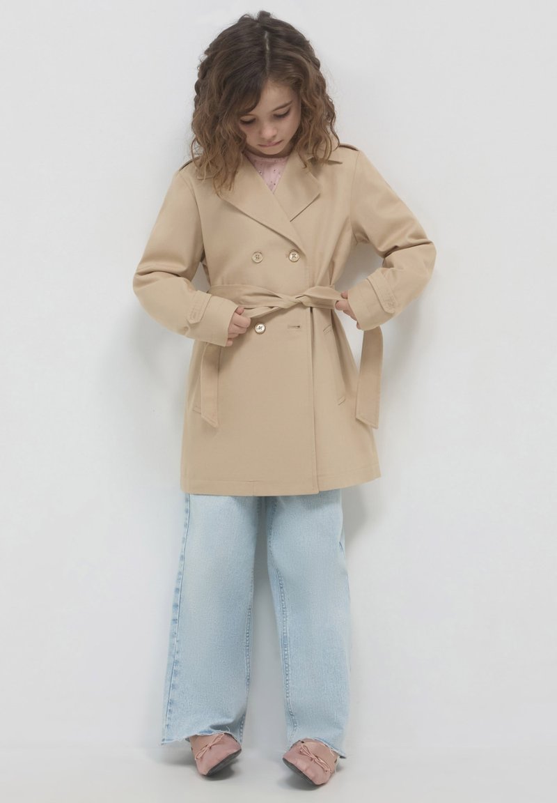 Beige Doppelreiher-Mantel mit Taillengürtel, langen Ärmeln und glatter Textur, kombiniert mit hellblauen, abgeriebenen Jeans und rosa Schuhen.