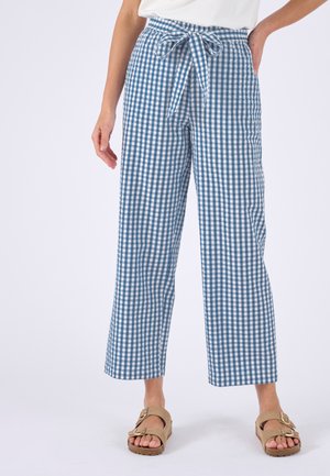 Vrouw draagt blauw-witte geruite wijde broek met een gestrikte tailleband en beige sandalen met gesp op een witte achtergrond.