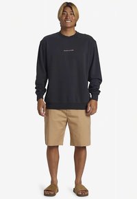 Navy sweatshirt with long sleeves and ribbed cuffs, featuring a small red logo at the chest; paired with tan shorts and brown slide sandals.