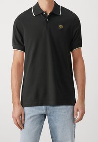 T-shirt polo noir avec un col et des bords de manches blancs contrastants, présentant un patch logo doré sur la poitrine. Matière en coton, coupe classique.