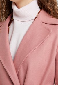 Růžový na míru šitý kabát přes světlý rolákový svetr. Kabát má výstřih s klopami a měkkou texturu, zatímco svetr je pruhovaný.