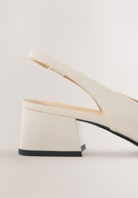 Escarpin slingback blanc avec talon carré bloc et bord de semelle noir sur fond clair.
