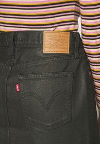 Poche arrière d'un jean Levi’s noir avec étiquette en cuir marron Levi Strauss et personne portant une chemise à manches longues rayée jaune, rose et marron.