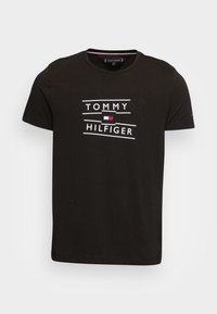 Camiseta negra de Tommy Hilfiger con texto de logo en blanco y pequeño emblema de bandera roja, blanca y azul centrado en el pecho, manga corta, cuello redondo.