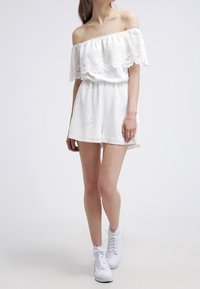 Romper bianco off-shoulder con sovrapposizione in pizzo testurizzato, vita elastica e vestibilità ampia. Abbinato a sneakers alte bianche.