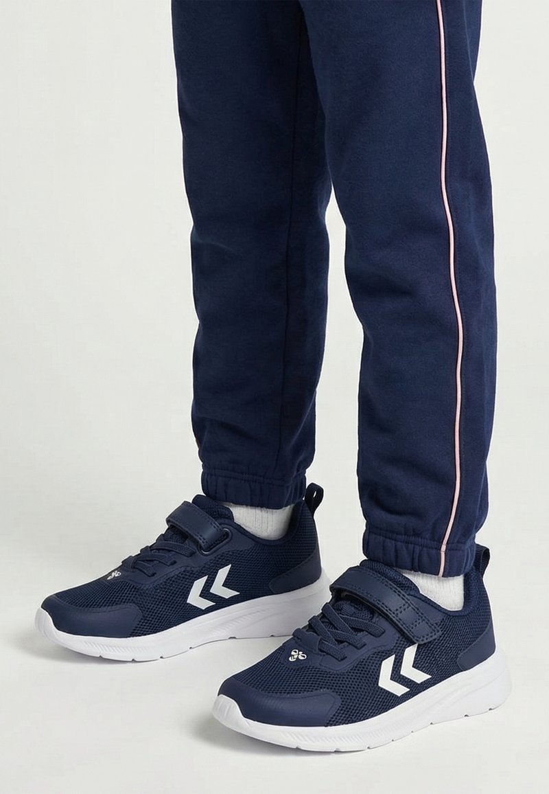 Kind trägt marineblaue Sneakers mit weißen Sohlen, Doppelchevron-Logo, Klettverschluss und marineblaue Jogginghose mit pinkem Seitenstreifen.