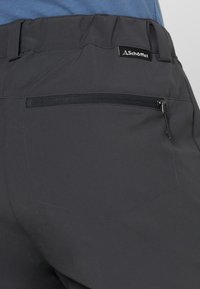 Pantalones gris oscuro con un bolsillo trasero con cremallera y una etiqueta de logo. La tela parece suave con un ligero brillo, lo que resalta su durabilidad.