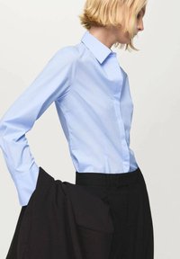 Svetlomodrá košeľa s dlhým rukávom a klasickým golierom, vyrobená z hladkej látky, v kombinácii s čiernymi nohavicami, predstavujúca formálne oblečenie.