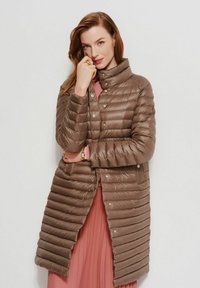 Brązowa kurtka puchowa z wysokim kołnierzem, z pikowanym wzorem, z zapięciem na napy oraz rozszerzanym krojem, noszona na różowej sukience.