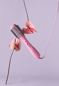 Brosse de coiffure rose avec des poils arrondis et un manche texturé, dotée d'un long cordon. L'arrière-plan est d'une douce couleur lavande avec des fleurs.