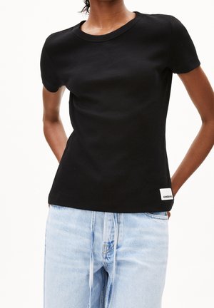 Personne portant un t-shirt noir uni à manches courtes et un jean taille haute bleu clair, debout les mains derrière le dos.