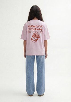 Persona con lunghi capelli scuri che indossa una maglietta rosa con testo marrone su un incontro sociale per un caffè domenicale e jeans azzurri a gamba larga.