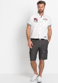 Camicia bianca a maniche corte con bottoni, polsini a scacchi neri e dettagli stampati, abbinata a pantaloni cargo grigi e sneaker bianche.