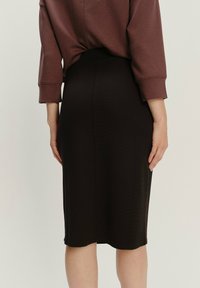Crna olovka suknja od glatke tkanine, uskog kroja, srednje dužine, s uskim pojasom, prikazana sa stražnje strane uz taupe duksericu.