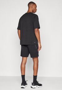 Férfi áll háttal, fekete bő pólót, cipzáras zsebű fekete rövidnadrágot, fekete zoknit és fekete-fehér sportcipőt visel.