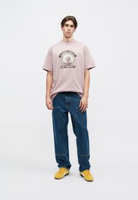 Roza bombažna majica s grafikonskim motivom slona in napisom "Carhartt Detroit", v kombinaciji z ohlapnimi modrimi kavbojkami in rumenimi supergami.