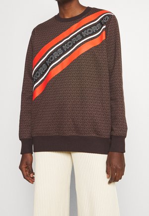Pull marron à motifs avec des rayures diagonales rouges, noires et blanches, ornées du texte répété "KORS", porté avec un pantalon côtelé écru.