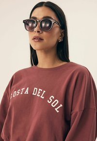 Bordowa bluza z napisem "COSTA DEL SOL" w białym kolorze, w połączeniu z okrągłymi, ciemnymi okularami przeciwsłonecznymi. Elegancka oprawka z błyszczącym wykończeniem.