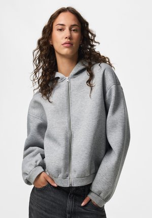 Kvinde med krøllet hår iført en lysegrå hættetrøje med lynlås og mørke jeans, stående foran en ensfarvet hvid baggrund.