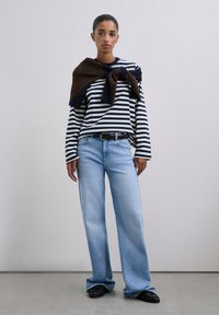 Camisa de rayas de manga larga en azul marino y blanco, combinada con pantalones vaqueros de pierna ancha en azul claro. Suéter marrón atado alrededor de los hombros; zapatos negros.