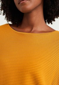 Woman with curly hair wearing a mustard yellow ribbed boatneck top, shown from neck to upper chest against a light background.