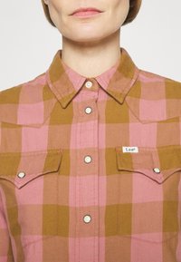 Femme portant une chemise à carreaux marron et rose boutonnée avec des poches à rabat, montrant le col et le haut de la poitrine.
