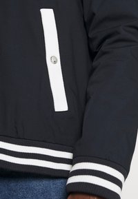 Veste bomber noire avec des poignets et des bordures de poche en côtes rayées blanches. Détail de bouton-pression en métal sur une bande verticale blanche. Tissu texturé doux.