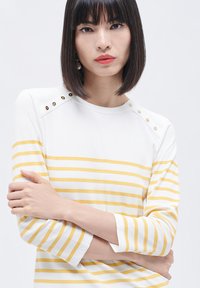 Witte en gele gestreepte top met driekwartmouwen, voorzien van decoratieve knopen op de schouders. Zachte, ribbelige stoftextuur.