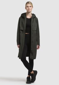 Long, dark green waterproof coat with a hood, front buttons, and two large pockets. Worn over black athletic attire and black sneakers.