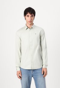 Cămașă verde deschis cu mânecă lungă și guler cu nasturi, având un mic logo pe piept, asortată cu blugi din denim albastru.