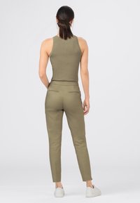 Žena stojící zády k fotoaparátu, oblečená v olivově zeleném bezrukávovém žebrovaném topu, sladěných úzkých kalhotách a bílých teniskách na jednolitém pozadí.