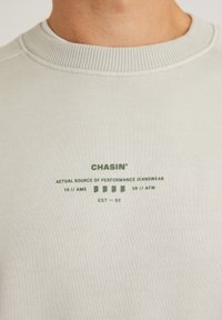 Sudadera de cuello redondo en gris claro, hecha de tela suave. Presenta texto impreso en verde, que incluye el nombre de la marca y varios detalles. Cuello de canalé.
