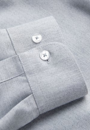 Puño de camisa de tela gris claro con dos botones de plástico transparente y costuras visibles sobre un material de textura suave.