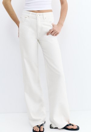 Personne portant un jean blanc taille haute à jambes larges et des tongs noires, avec une main dans la poche sur un fond blanc uni.
