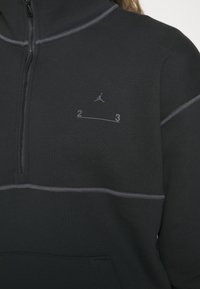 Sudadera polar negra con cuello alto, cremallera hasta un cuarto, costuras grises y logotipo de Jordan. Presenta una línea horizontal con el texto "2 3" debajo.