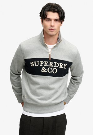 Junger Mann mit dunklen Haaren, der einen grauen Sweatshirt mit Viertel-Reißverschluss trägt, mit dem Text "SUPERDRY & GO" auf einem schwarzen Panel über der Brust.