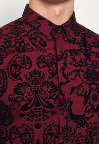 Viininpunainen paita, jossa musta damaskikuvio, nappikiinnitteinen kaulus ja teksturoitu kangas, esittelee monimutkaisia kukka- ja geometrisia design-elementtejä.