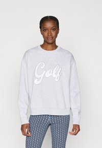 Sweatshirt cinza com 'Golf' bordado em branco em letra cursiva. Apresenta um decote redondo e mangas longas. Combinado com leggings azuis estampadas.
