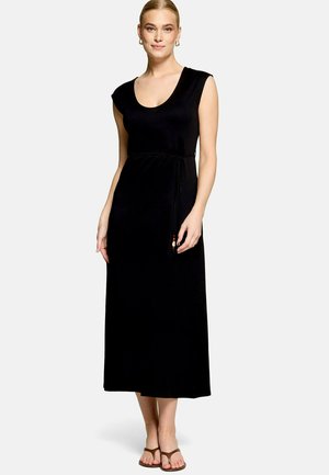 Femme portant une robe midi noire sans manches avec un décolleté arrondi et une taille nouée, associée à des sandales marron et des boucles d'oreilles créoles dorées.