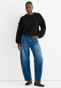 Black textured sweater with puffed details, paired with light blue high-waisted jeans and black flat shoes, against a white background.