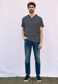 Raidallinen lyhythihainen henley-paita tummansinisessä ja valkoisessa, yhdistettynä sinisiin farkkuihin. Malli, joka pitää päällään tummansiniset lenkkarit, seisoo yksinkertaisen taustan edessä.