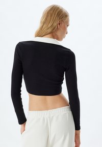Musta ribbed pitkähihaisen crop top, jossa on valkoinen kaulus, esittelee tiukkaa muotoilua. Mallilla on päällään vaaleanväriset, löysät housut.