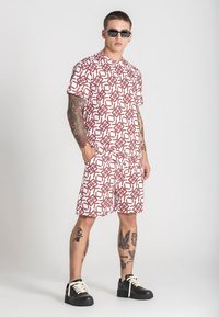 Bijela majica kratkih rukava i šorts s crvenim geometrijskim uzorkom. Materijal djeluje lagano. Kombinirano s crnim tenisicama i sunčanim naočalama.