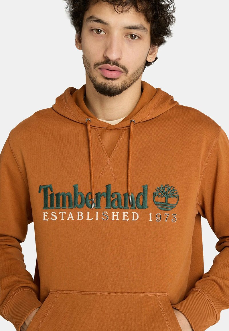 Sweat à capuche orange avec un logo à l'avant en texte vert indiquant "Timberland ÉTABLI EN 1973" et un emblème d'arbre, fabriqué à partir d'un tissu doux.