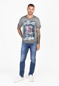 Camiseta de algodón gris con estampado gráfico de motocicleta desgastado. Combinada con jeans de mezclilla azules y zapatillas blancas. Ajuste casual y mangas cortas.